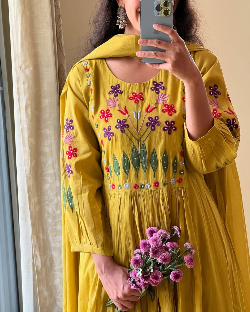 Person wearing a yellow embroidered outfit holding pink flowers, taking a mirror selfie.